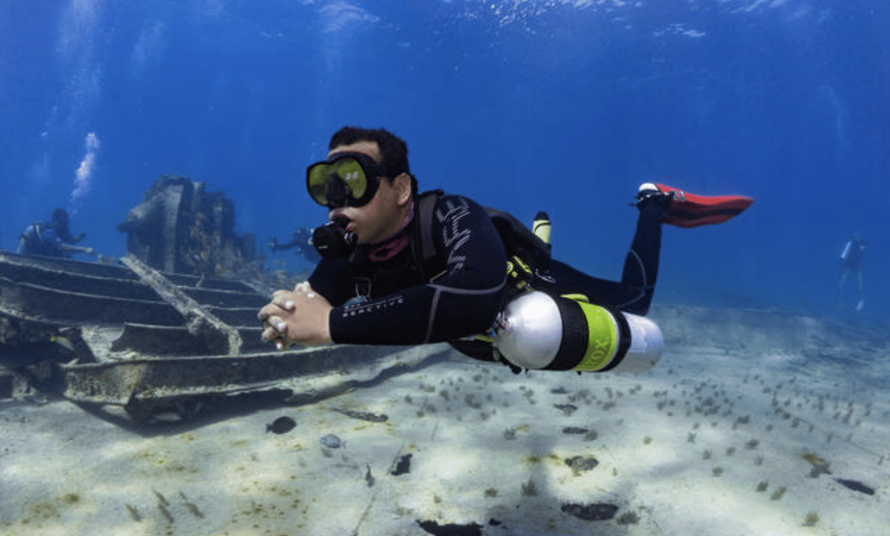 Screenshot 2026-04-01 at 221610 - N9BO℠ | Global Underwater Services Ltd A scuba diver in a wetsuit and diving gear swims near a sunken shipwreck on the ocean floor, with other divers visible in the background and sunlight filtering through the clear blue water.
