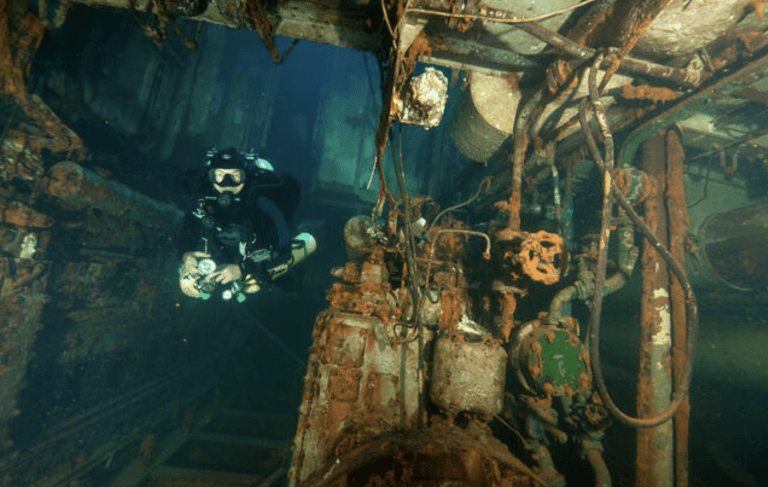 A scuba diver explores the rusted interior of a sunken shipwreck, surrounded by corroded pipes, machinery, and debris in the underwater environment.