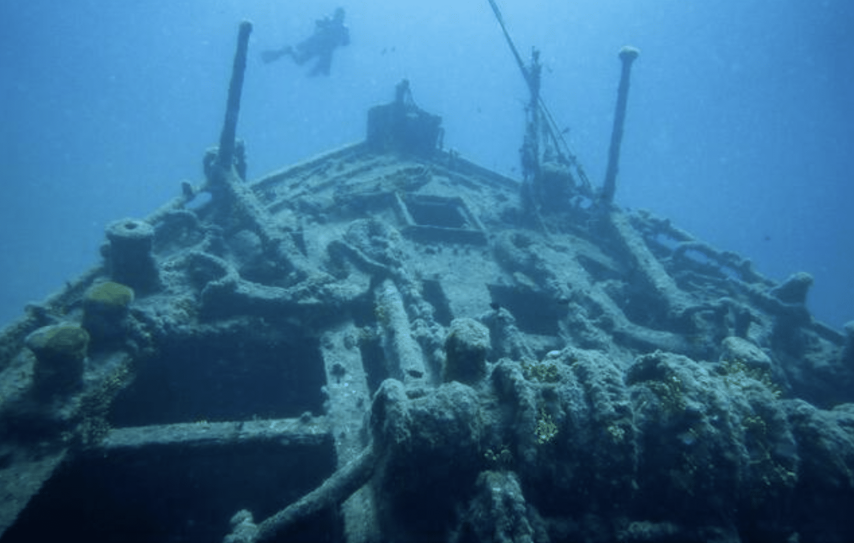 Screenshot 2026-04-01 at 225145 - N9BO℠ | Global Underwater Services Ltd A sunken shipwreck lies on the ocean floor, covered in marine growth and corals, with a scuba diver swimming above it in the blue water.