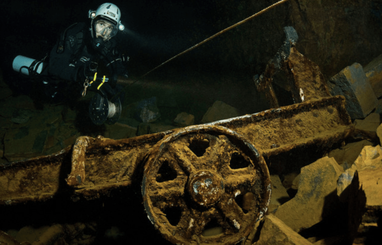 A scuba diver in full kit explores a dark underwater scene with rocky debris and a large, rusted metal wheel in the foreground.