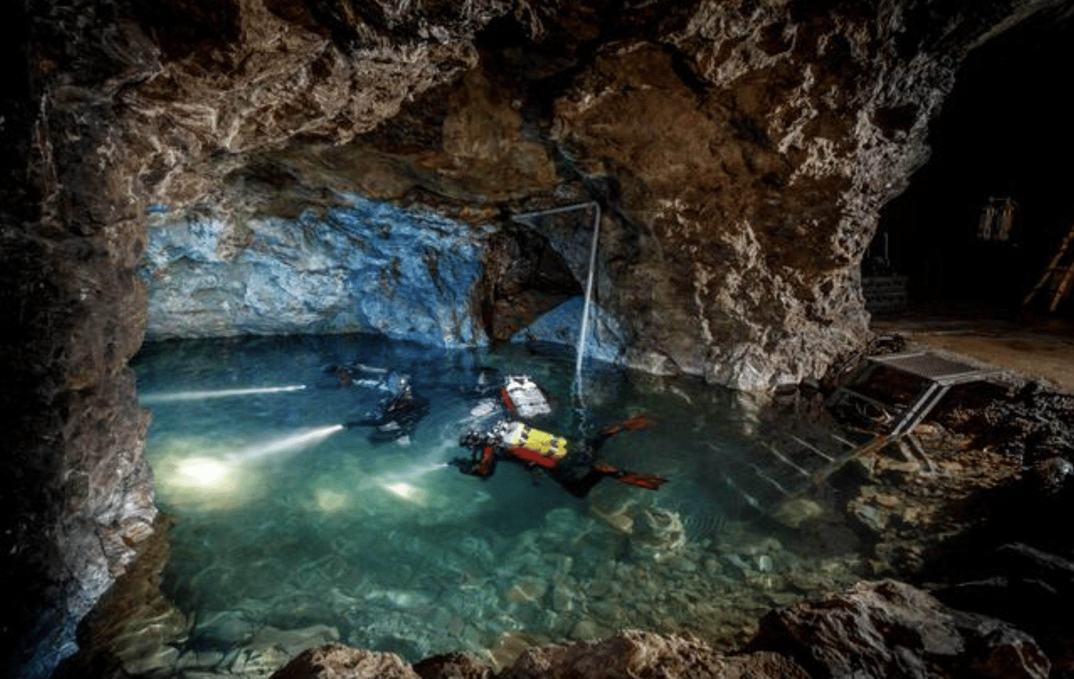 Screenshot 2026-04-01 at 232508 - N9BO℠ | Global Underwater Services Ltd Two scuba divers with torches explore a clear, rocky underground cave pool. Metal stairs and railings lead into the water from the cave floor, illuminated by the divers’ lights and some ambient cave lighting.