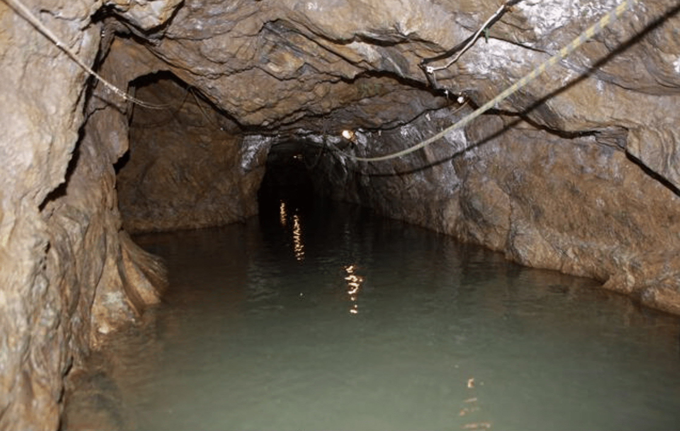 Screenshot 2026-04-01 at 232526 - N9BO℠ | Global Underwater Services Ltd Flooded tunnel inside a rocky cave or mine, with rough stone walls and ceiling, partially submerged in water, and electrical wires hanging along the sides.