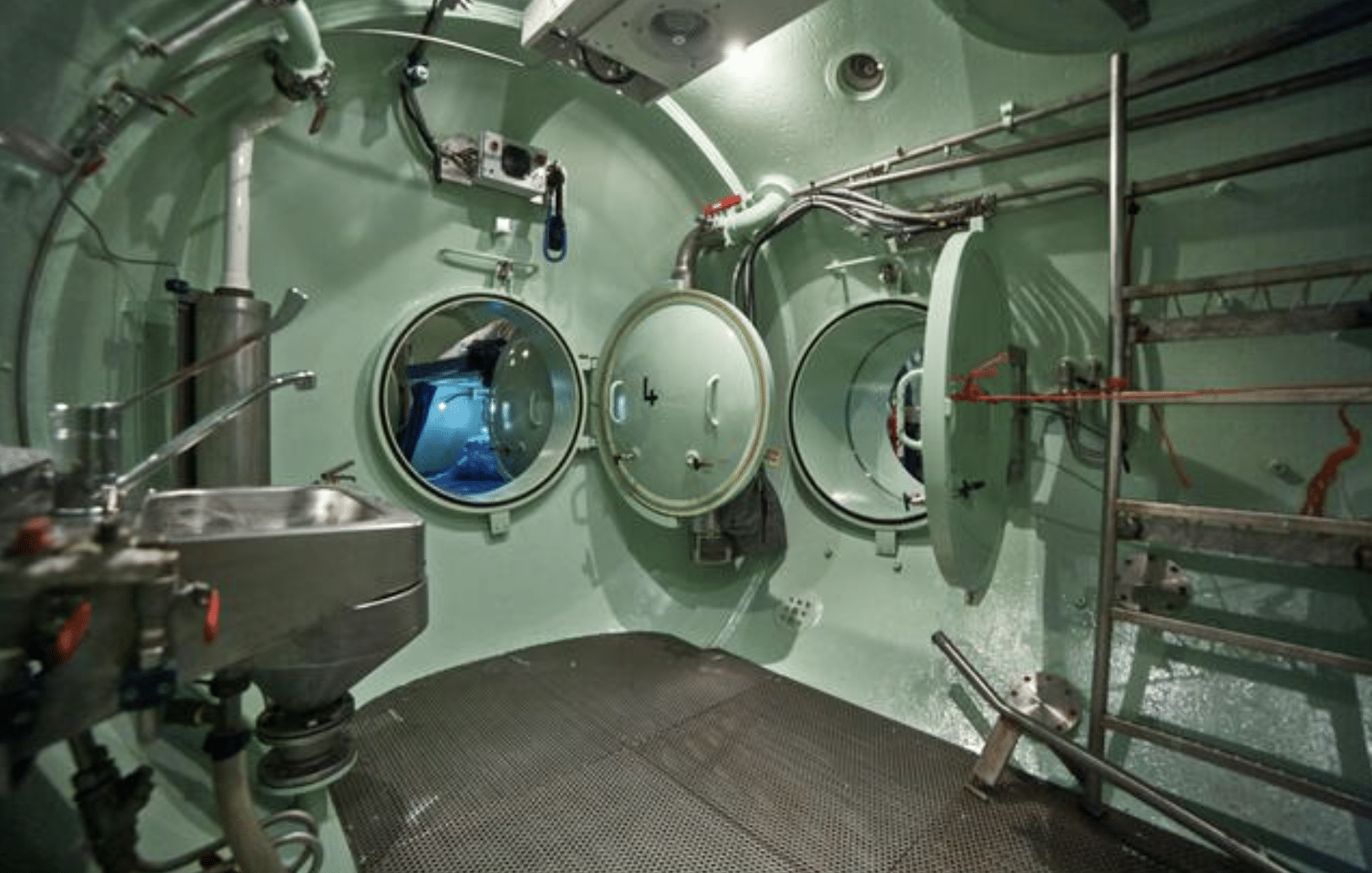 Screenshot 2026-04-01 at 234818 - N9BO℠ | Global Underwater Services Ltd Interior of a pressurised chamber with round airtight doors, metal floor, a sink, pipes, and various equipment. The chamber has a clinical, industrial appearance and is illuminated by overhead lighting.