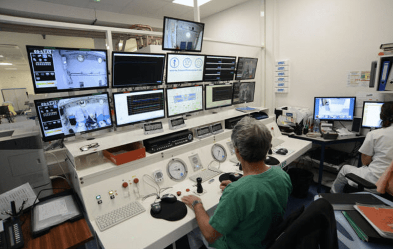 A person in medical scrubs sits at a control desk with multiple monitors, gauges, and control panels in a room, whilst another person works at a computer in the background.