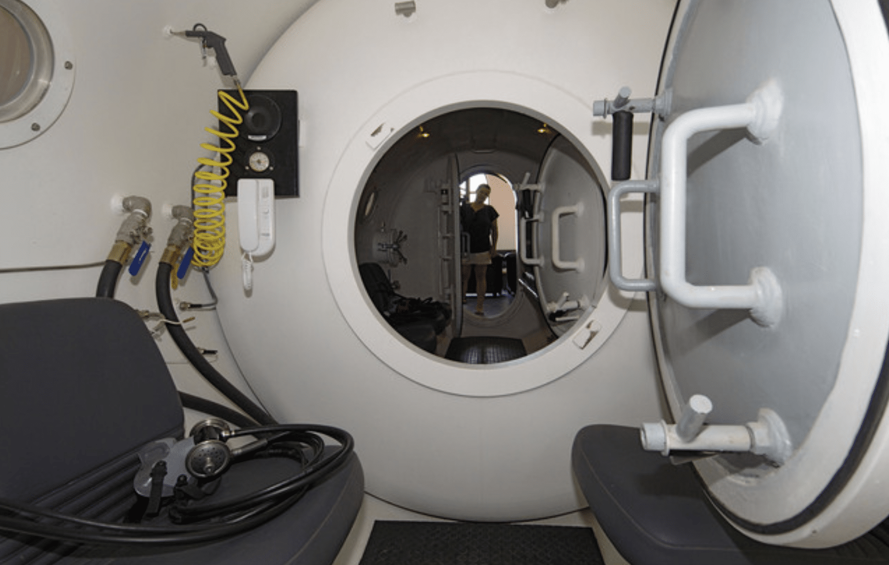Screenshot 2026-04-01 at 234846 - N9BO℠ | Global Underwater Services Ltd Interior view of a hyperbaric chamber with open hatch doors, two benches, medical equipment, and a person visible standing in the background through the circular doorway.