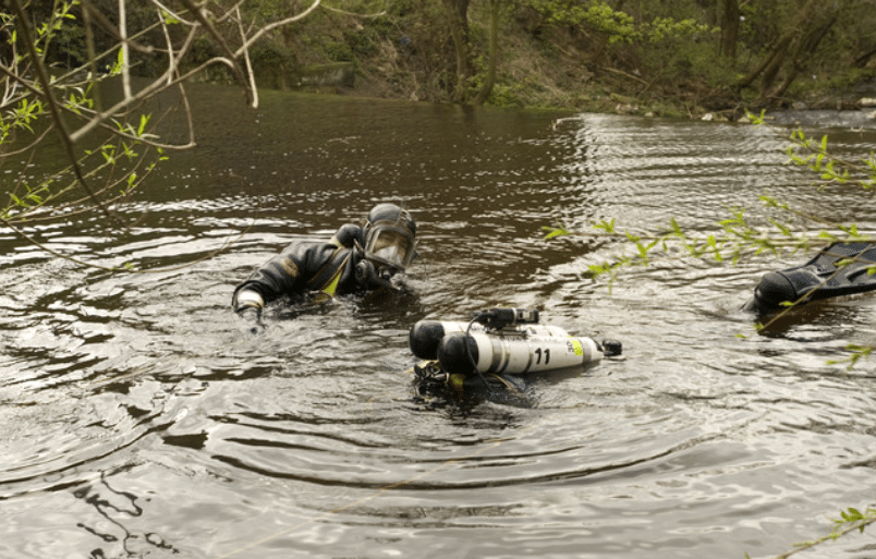 Screenshot 2026-04-02 124658 - N9BO℠ | Global Underwater Services Ltd Two scuba divers in wetsuits and masks are partially submerged in a calm, wooded river. One diver has an air cylinder labelled 21 floating nearby. Green branches frame the scene.