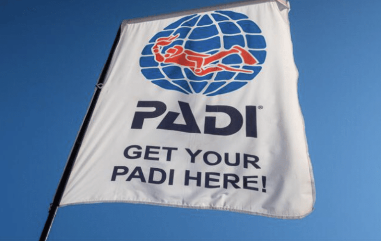 A white flag with the PADI logo, featuring a red scuba diver over a blue globe, and the text PADI GET YOUR PADI HERE! against a clear blue sky.