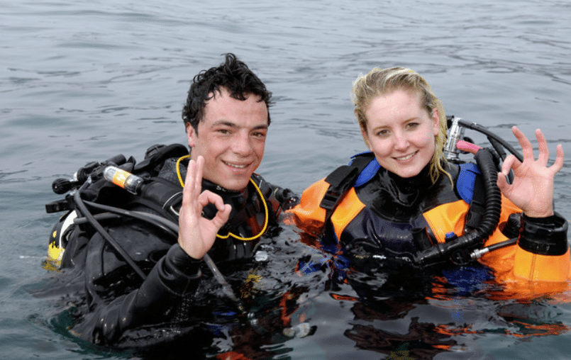 Screenshot 2026-04-02 131118 - N9BO℠ | Global Underwater Services Ltd Two scuba divers wearing wetsuits and gear are in the water, smiling, and making the “OK” hand sign with one hand each. The water around them appears calm.