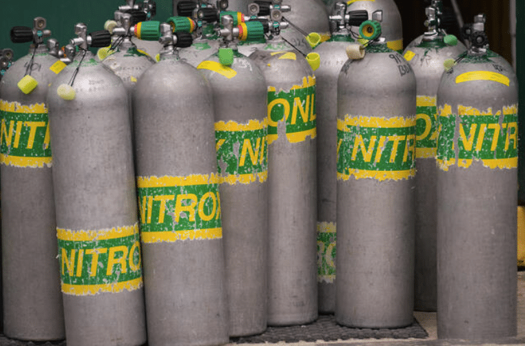 Several large metal scuba cylinders with worn yellow and green “NITROX” labels are standing upright and grouped closely together, ready for use, against a green background.