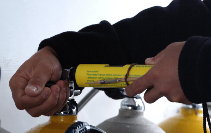 Screenshot 2026-04-02 132819 - N9BO℠ | Global Underwater Services Ltd Close-up of a person’s hands attaching a yellow air tank or oxygen cylinder, possibly for diving or safety purposes, with a black sleeve visible on one arm.