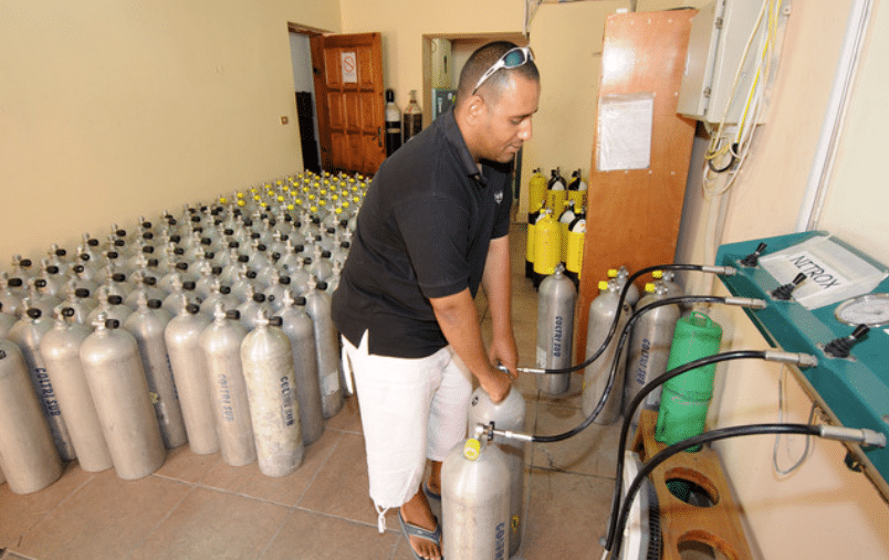 Screenshot 2026-04-02 133025 - N9BO℠ | Global Underwater Services Ltd A man in a black shirt and white shorts fills a metal scuba cylinder with air in a room containing many similar cylinders lined up against the wall.