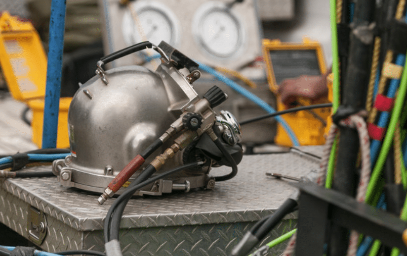 Screenshot 2026-04-02 151243 - N9BO℠ | Global Underwater Services Ltd A close-up of a metal diving helmet with attached hoses resting on a metal surface, surrounded by colourful cables and equipment, with blurred gauges and a person in the background.