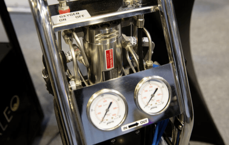 Close-up of a metal machine with two pressure gauges labelled IN and OUT, various knobs, tubes, and a switch for oxygen marked ON and OFF. The device appears industrial or scientific.