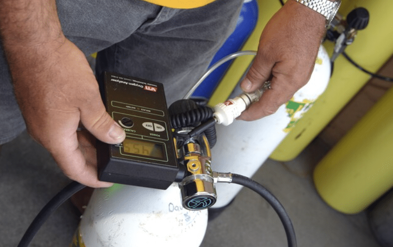 Screenshot 2026-04-02 153039 - N9BO℠ | Global Underwater Services Ltd A person uses an oxygen analyser device connected to a scuba cylinder, checking the oxygen concentration. Several other scuba cylinders are visible in the background.