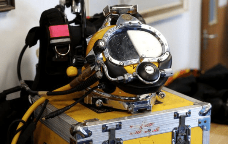 A yellow and black commercial diving helmet with attached breathing hoses is resting on a yellow equipment case, with a diving harness and other gear visible in the background.