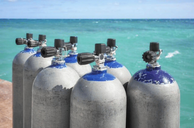 Screenshot 2026-04-02 154208 - N9BO℠ | Global Underwater Services Ltd A group of scuba diving air cylinders with blue tops arranged on a quay, positioned near turquoise sea water under a clear sky.