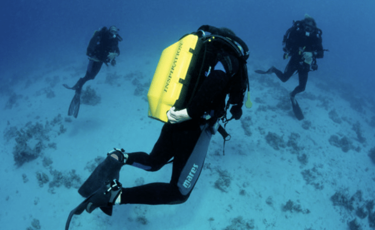 Three scuba divers swim underwater above a sandy, rocky sea floor. The diver in the foreground wears a yellow cylinder marked INSPIRATION, while two other divers are visible in the background.