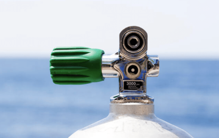A close-up of the valve on a scuba diving cylinder with a green handle, set against a blurred background of the sea and sky.