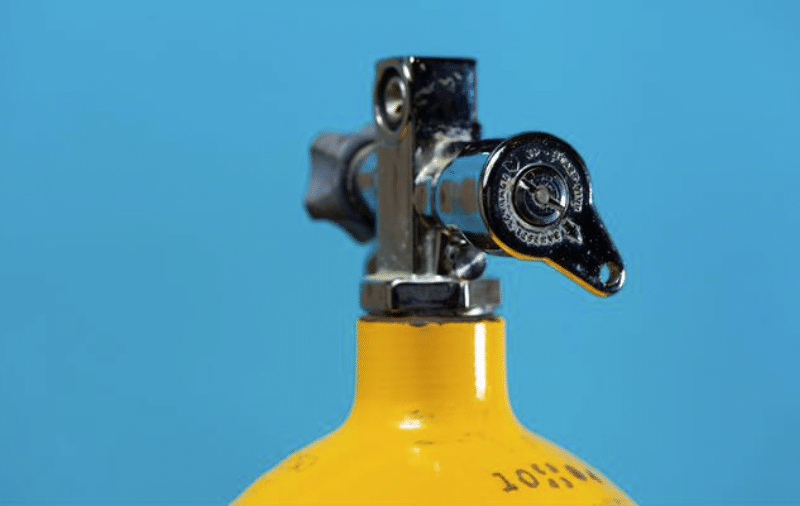 Screenshot 2026-04-03 151324 - N9BO℠ | Global Underwater Services Ltd A close-up of the valve and top portion of a yellow scuba diving cylinder against a solid blue background.