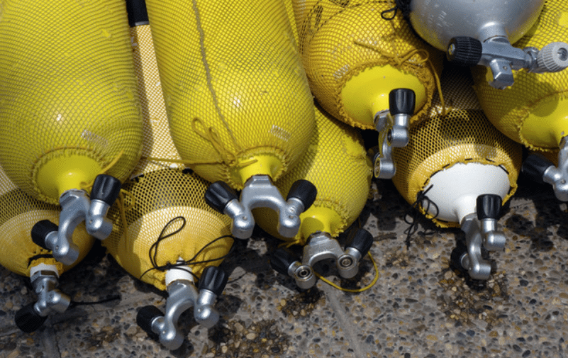 Screenshot 2026-04-03 151416 - N9BO℠ | Global Underwater Services Ltd A group of yellow scuba diving cylinders with protective mesh covers are lying on a rough, pebbled surface. The cylinders are arranged side by side, with their valves and regulators facing outward.