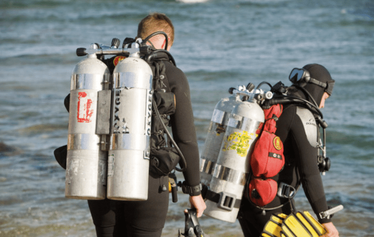 Two scuba divers in wetsuits, carrying large oxygen cylinders on their backs, walk towards the water from the shore. The sea appears calm, and the weather looks clear.