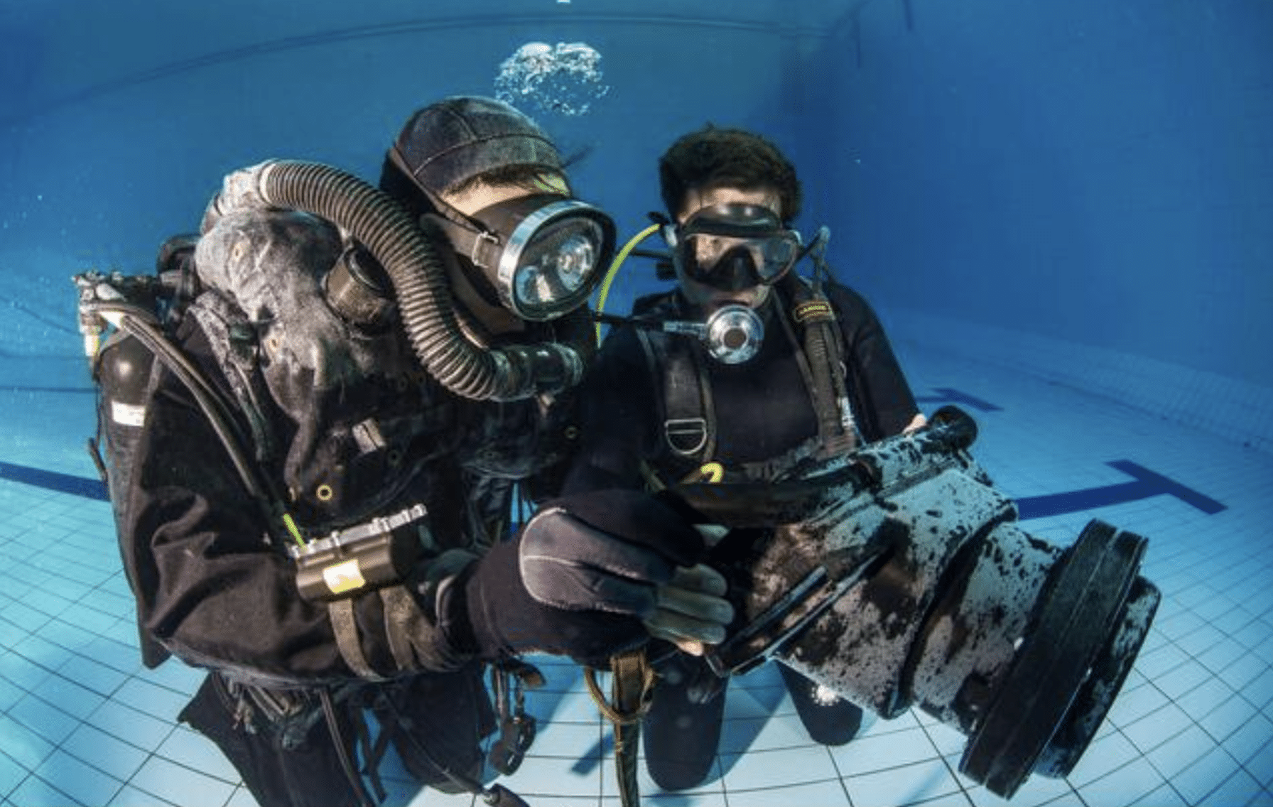 Two scuba divers in wetsuits and masks are underwater in a swimming pool, closely examining a large underwater camera housing whilst surrounded by blue water and tiled pool floor.