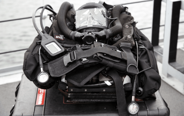 A close-up of a technical diving rebreather setup, including a full-face mask, hoses, control display, pressure gauges, and harness, resting on a black equipment case on a boat deck.