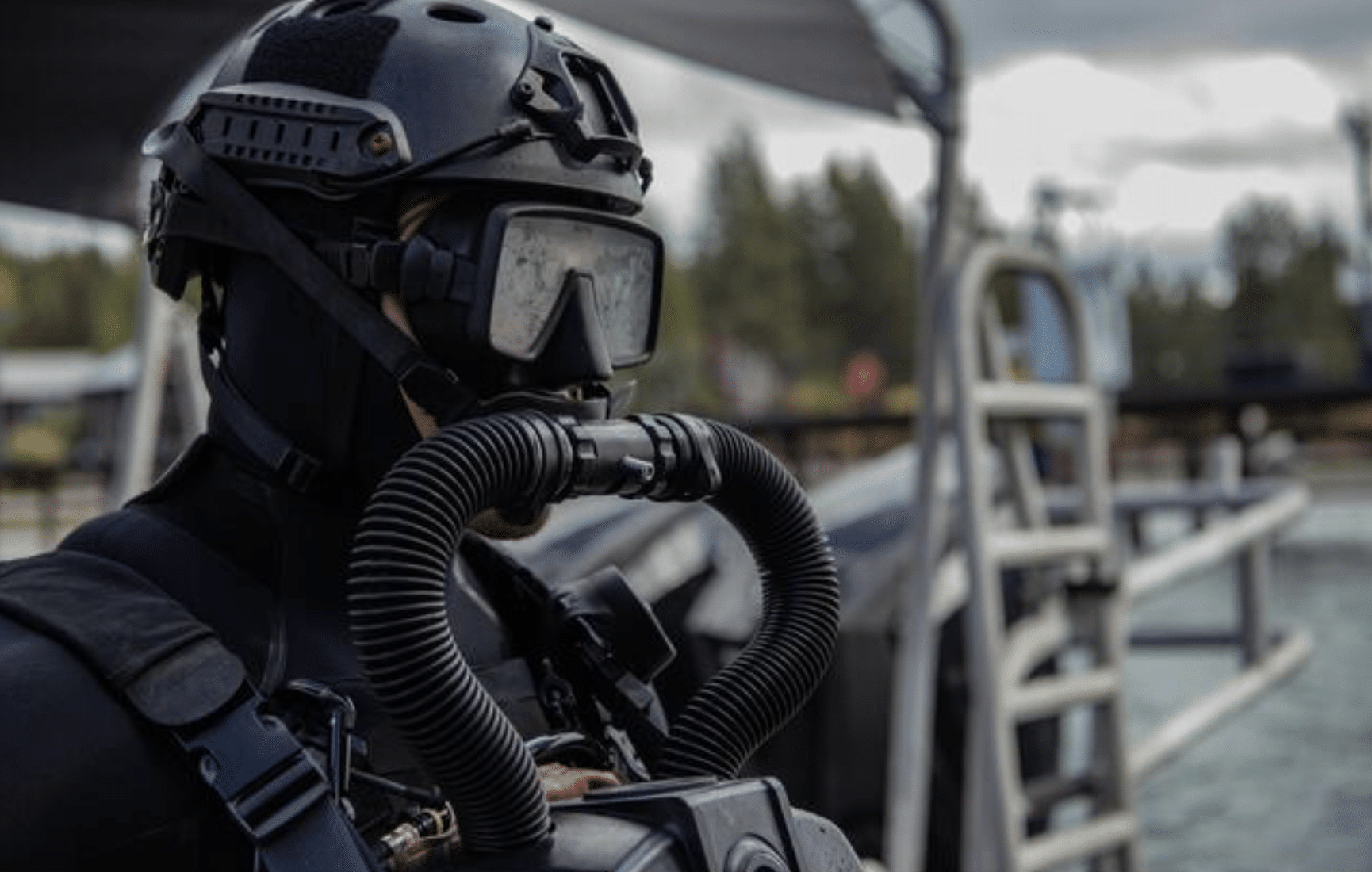 A diver in full black gear, including a helmet, mask, and rebreather, stands near water with metal railings and a ladder in the background. Trees and blurred outdoor elements are visible.