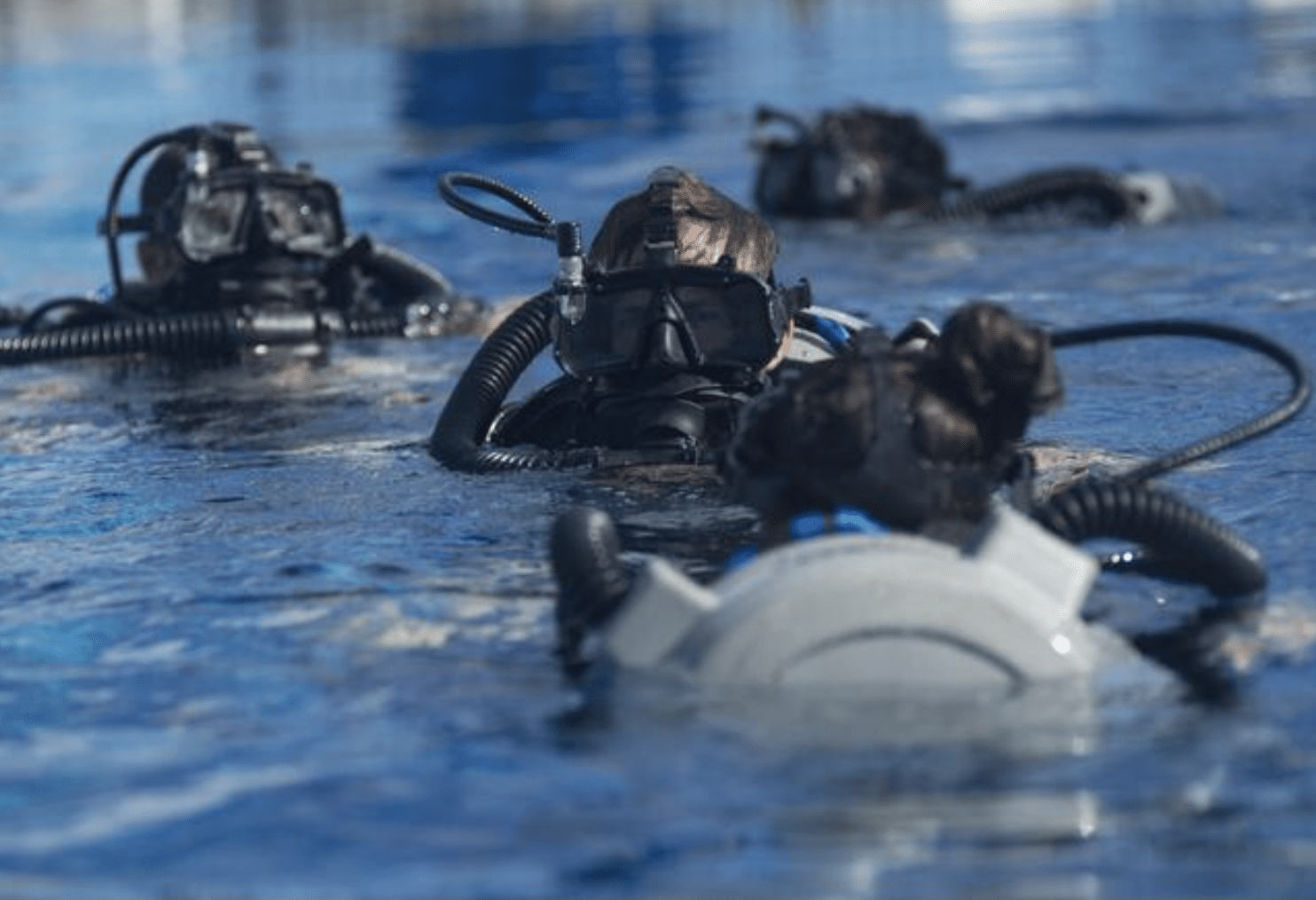 Four divers in black wetsuits and full-face masks swim in a body of water, wearing breathing apparatus with hoses visible above the surface. The image is focused on the diver in front.