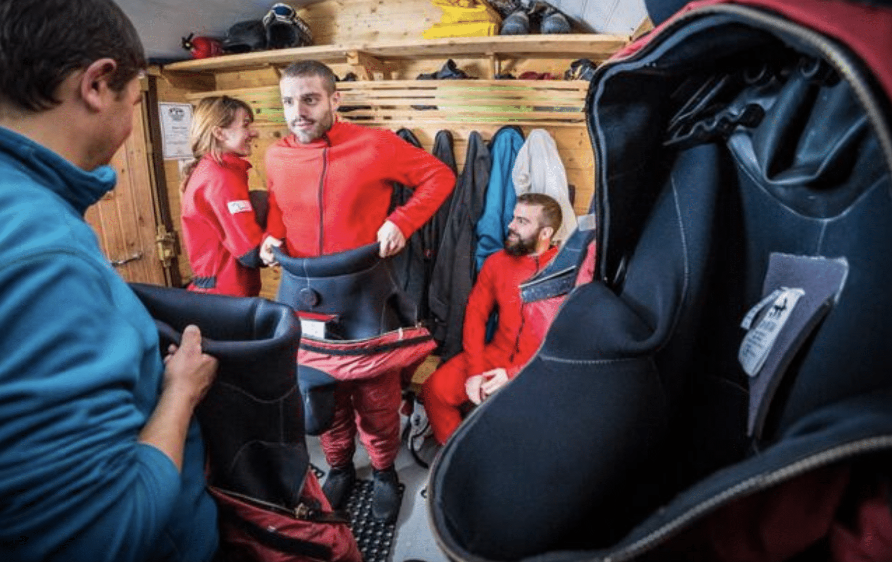 Screenshot 2026-04-05 at 174649 - N9BO℠ | Global Underwater Services Ltd Four people in a wooden room prepare for an activity, putting on red suits. Two stand while two sit, all focused on getting dressed. One person holds up a large suit in the foreground, partially out of focus.