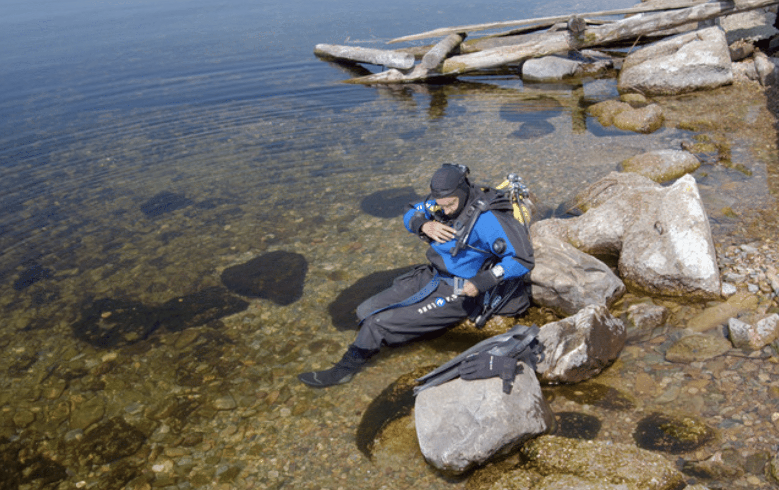 Screenshot 2026-04-05 at 174717 - N9BO℠ | Global Underwater Services Ltd A scuba diver in a blue drysuit sits on rocks at the edge of a clear, shallow lake, surrounded by large stones and driftwood, adjusting their kit with fins resting nearby.