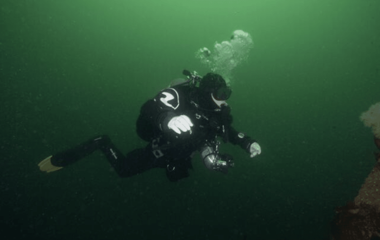 A scuba diver in a black wetsuit and yellow fins swims underwater in green-tinted water, releasing a stream of bubbles whilst holding an underwater camera.
