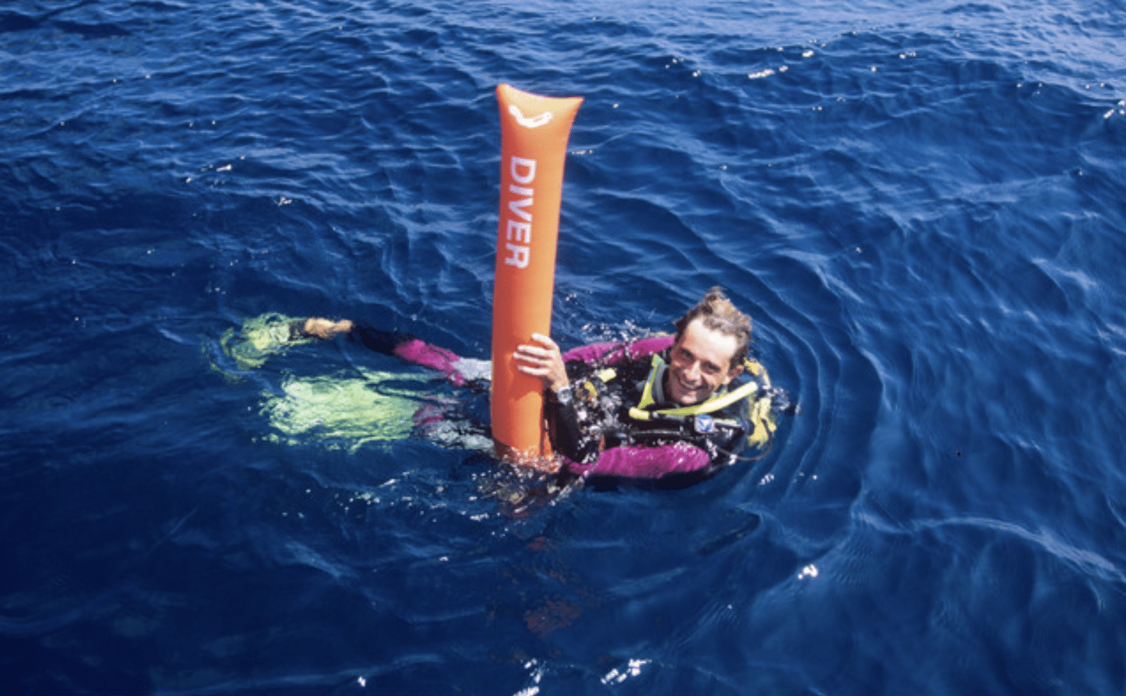 Screenshot 2026-04-05 at 180440 - N9BO℠ | Global Underwater Services Ltd A scuba diver floats in deep blue water, smiling and holding an orange inflatable signal tube labelled DIVER. The diver wears a wetsuit, scuba gear, and yellow fins.