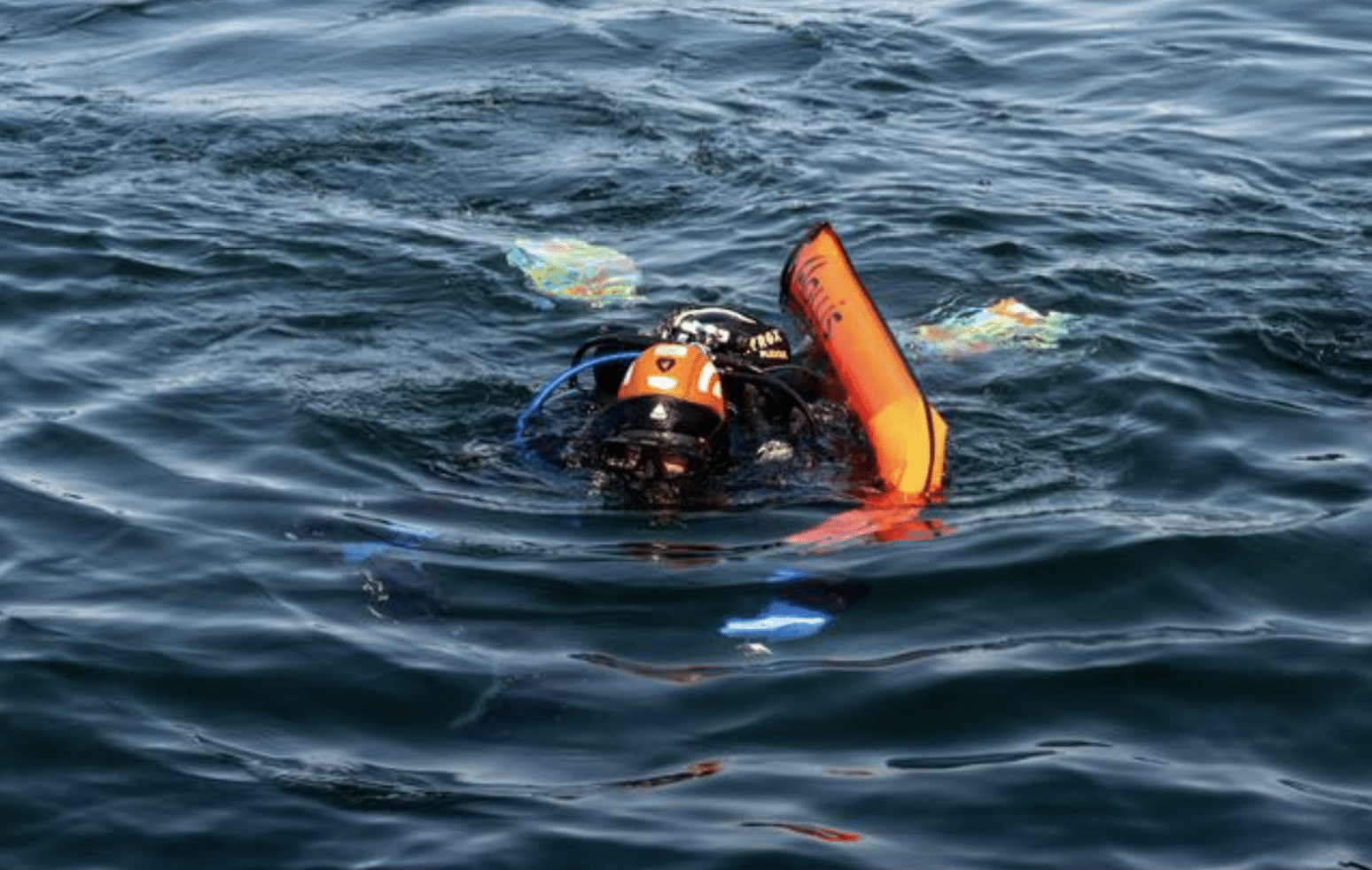 Screenshot 2026-04-05 at 180448 - N9BO℠ | Global Underwater Services Ltd A scuba diver wearing an orange helmet and carrying an orange flotation device floats on the surface of dark, rippling water. The diver's fins are visible beneath the water.
