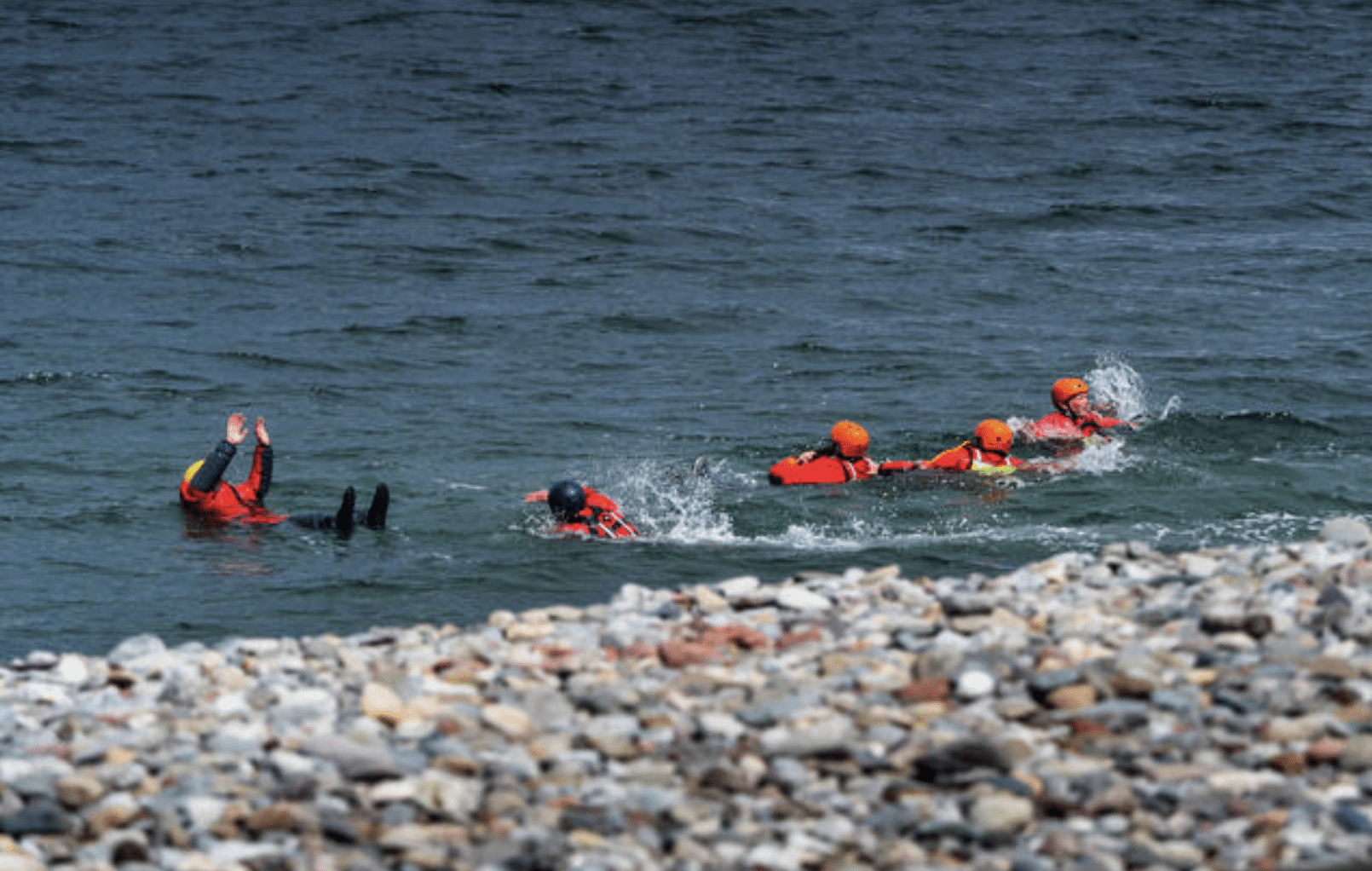 Screenshot 2026-04-05 at 181558 - N9BO℠ | Global Underwater Services Ltd Several people in red wetsuits and helmets are swimming in choppy water near a rocky shore, with one person slightly separated from the group, reaching their arms upwards.