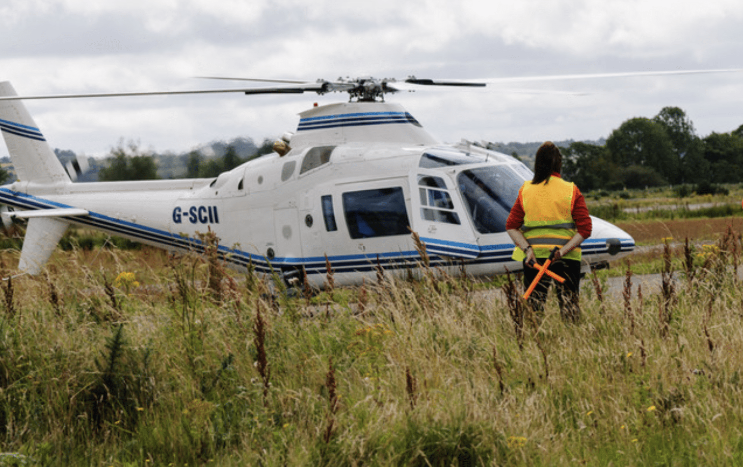 Screenshot 2026-04-05 at 190335 - N9BO℠ | Global Underwater Services Ltd A person in a yellow safety vest stands in front of a white helicopter with blue stripes, holding orange wands, on a grassy airfield with trees in the background.