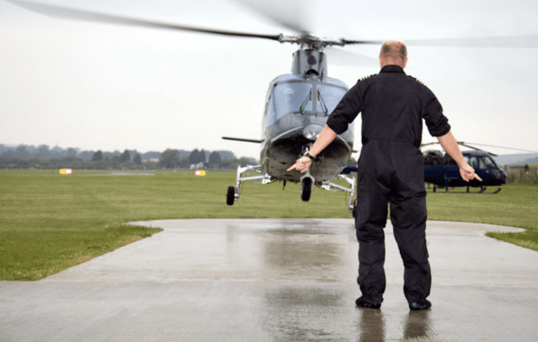 A person in a black boiler suit stands on a helipad, guiding a helicopter as it lands on a grassy field, with another helicopter and trees visible in the background.
