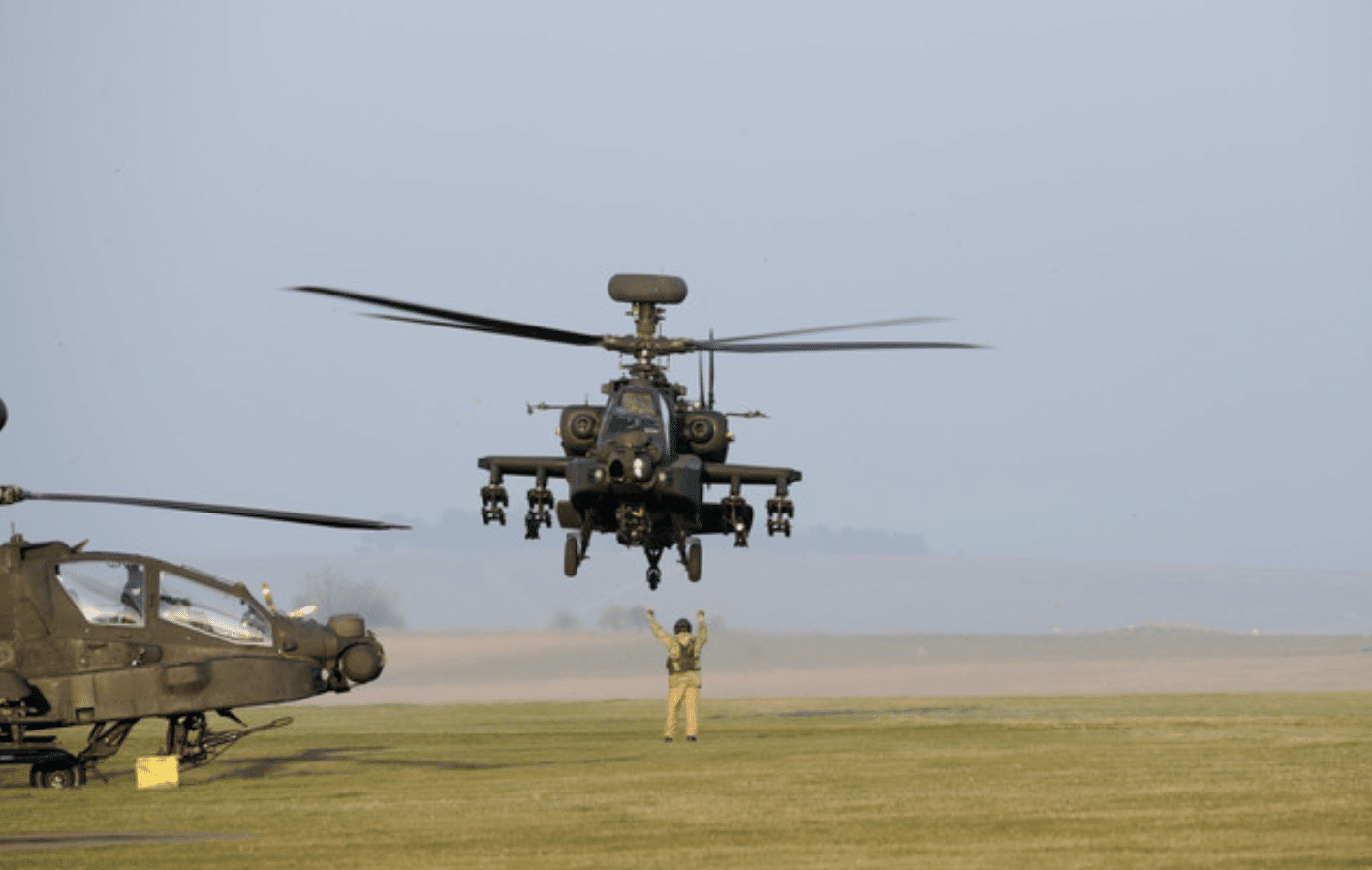 Screenshot 2026-04-05 at 190359 - N9BO℠ | Global Underwater Services Ltd A person on the ground signals as a military helicopter hovers above the grass, with another helicopter parked nearby and open fields in the background.