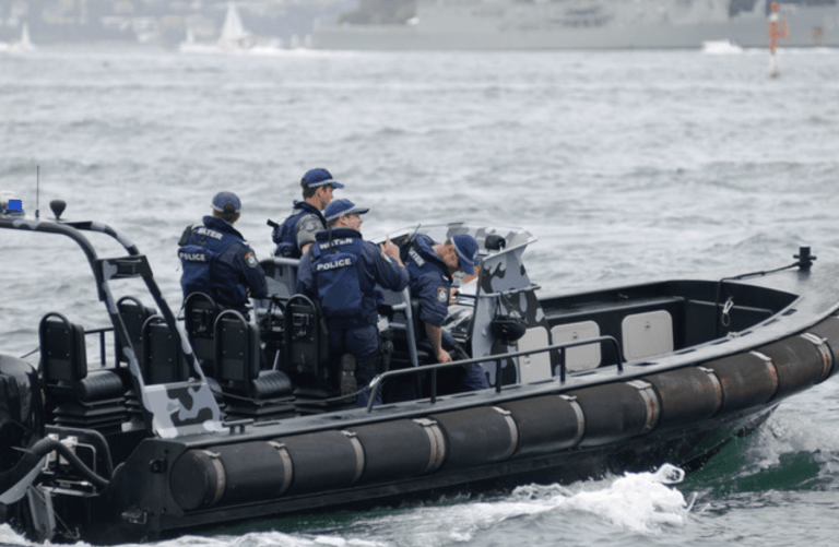 A group of police officers in uniform ride in a black inflatable patrol boat on the water. The boat has camouflage panels, and the officers appear focused as they navigate the choppy waves. A large ship is visible in the background.