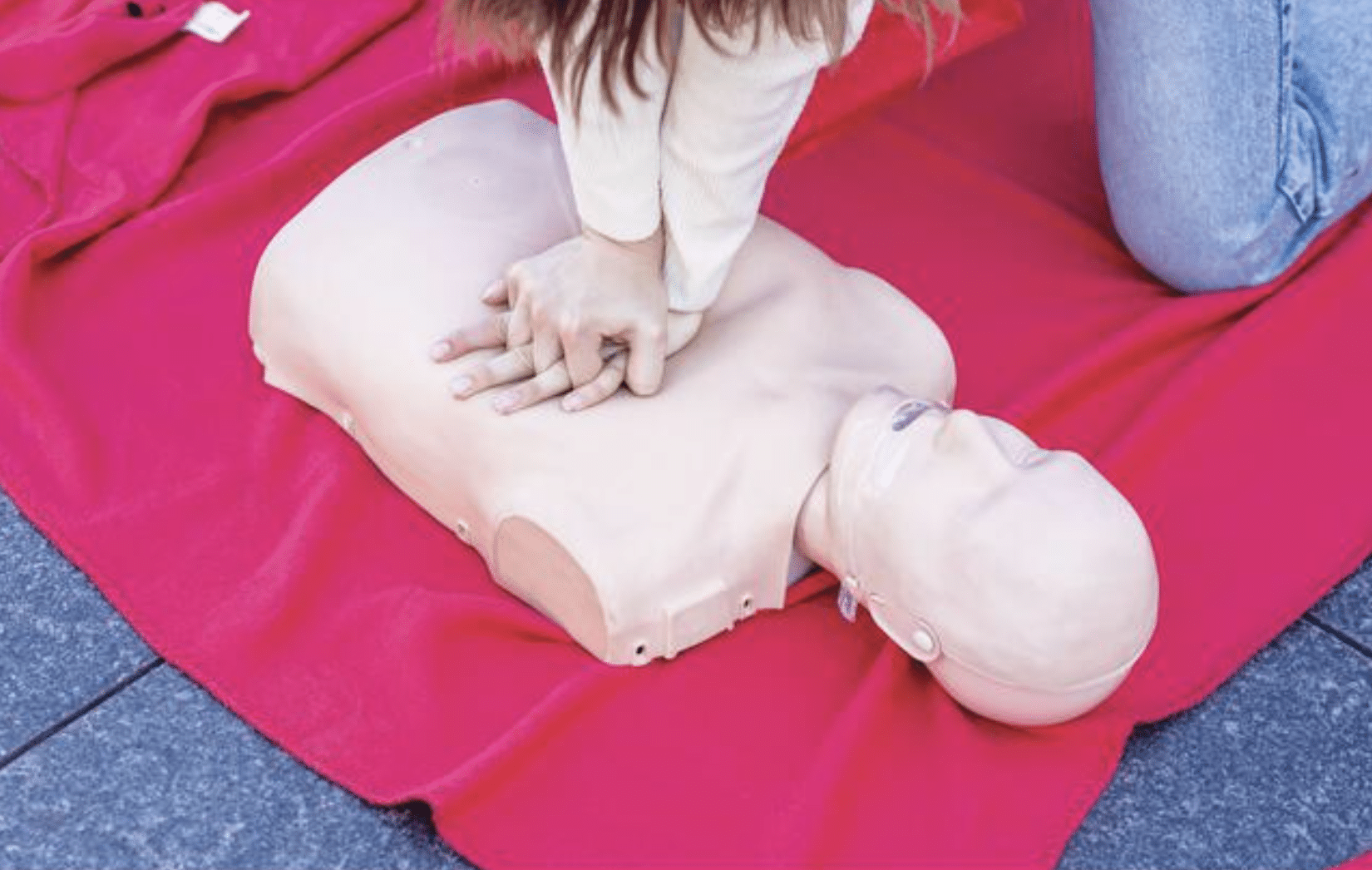 A person practises CPR by performing chest compressions on a training manikin placed on a red blanket.