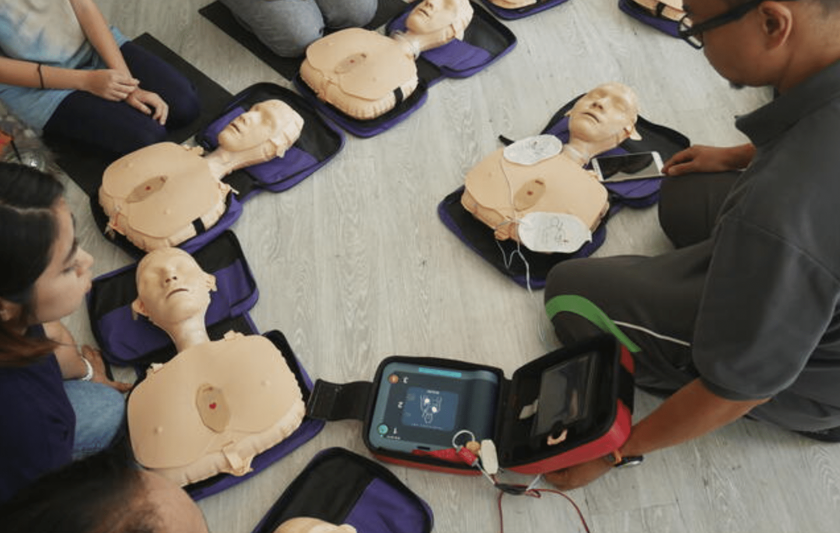 A group of people sit around CPR training manikins on the floor as an instructor demonstrates how to use an automated external defibrillator (AED) on one of the manikins.