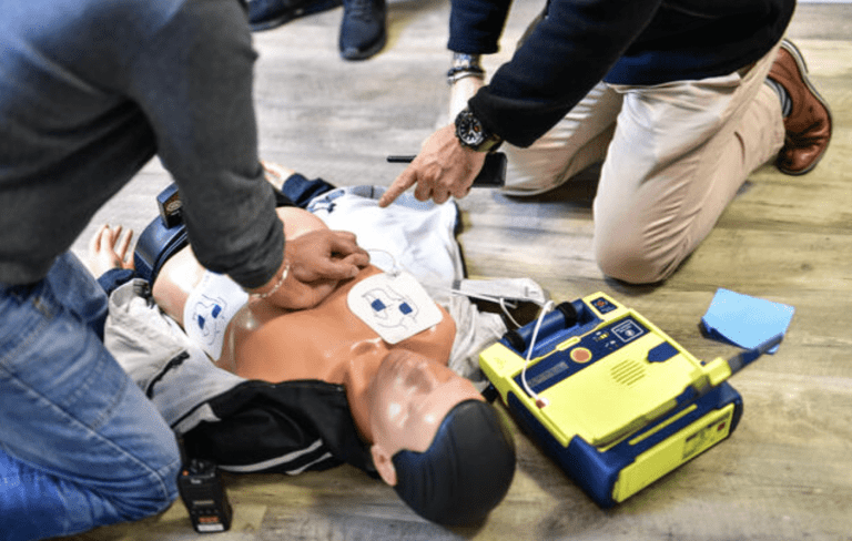 Two people kneel on the floor practising CPR and using an automated external defibrillator (AED) on a medical training manikin wearing a jacket. The AED is open and positioned next to the manikin.