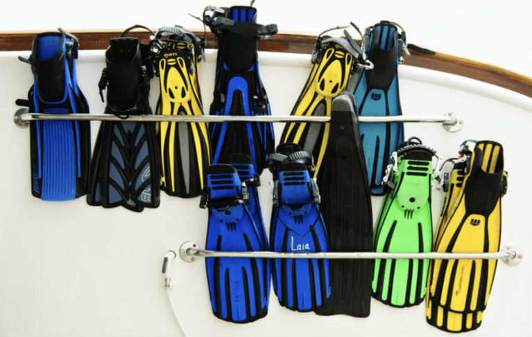 A variety of colourful snorkelling and diving fins in blue, black, yellow, and green are neatly arranged on two metal racks against a white wall.