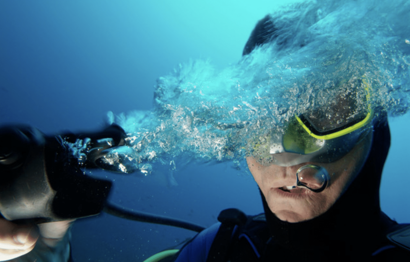 Screenshot 2026-04-05 at 224514 - N9BO℠ | Global Underwater Services Ltd A scuba diver wearing a wetsuit and mask releases air underwater, creating a burst of bubbles that partially obscures their face. The background is a deep blue, indicating they are submerged in the sea.
