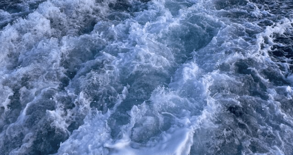 Churning ocean water creates foamy white waves and swirling patterns, likely from the wake of a boat moving through deep blue water.
