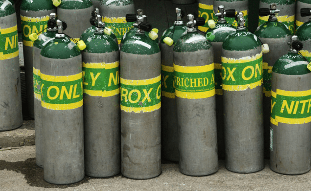 A group of silver and green metal gas cylinders with yellow and green labels, standing upright on a concrete surface. The labels read “ENRICHED AIR NITROX ONLY”.