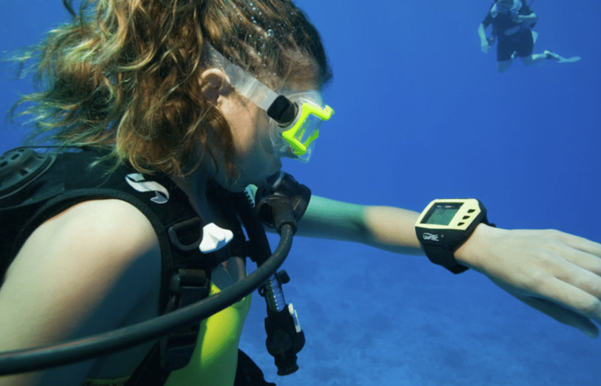 A scuba diver underwater checks a yellow dive computer on their wrist. The diver wears a mask, snorkel, and scuba gear, with another diver swimming in the background against the blue sea.