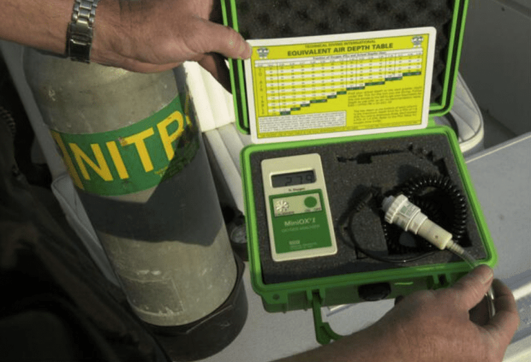 A person holds a green case containing a MiniOX I oxygen analyser and a chart. Beside the case, there is a scuba cylinder labelled NITROX. The case is foam-lined to protect the analyser.