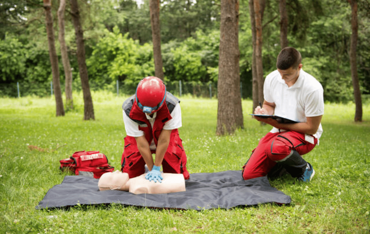A person in safety gear practises CPR on a training dummy outdoors while another person kneels nearby, taking notes on a clipboard. They are surrounded by grass and trees.