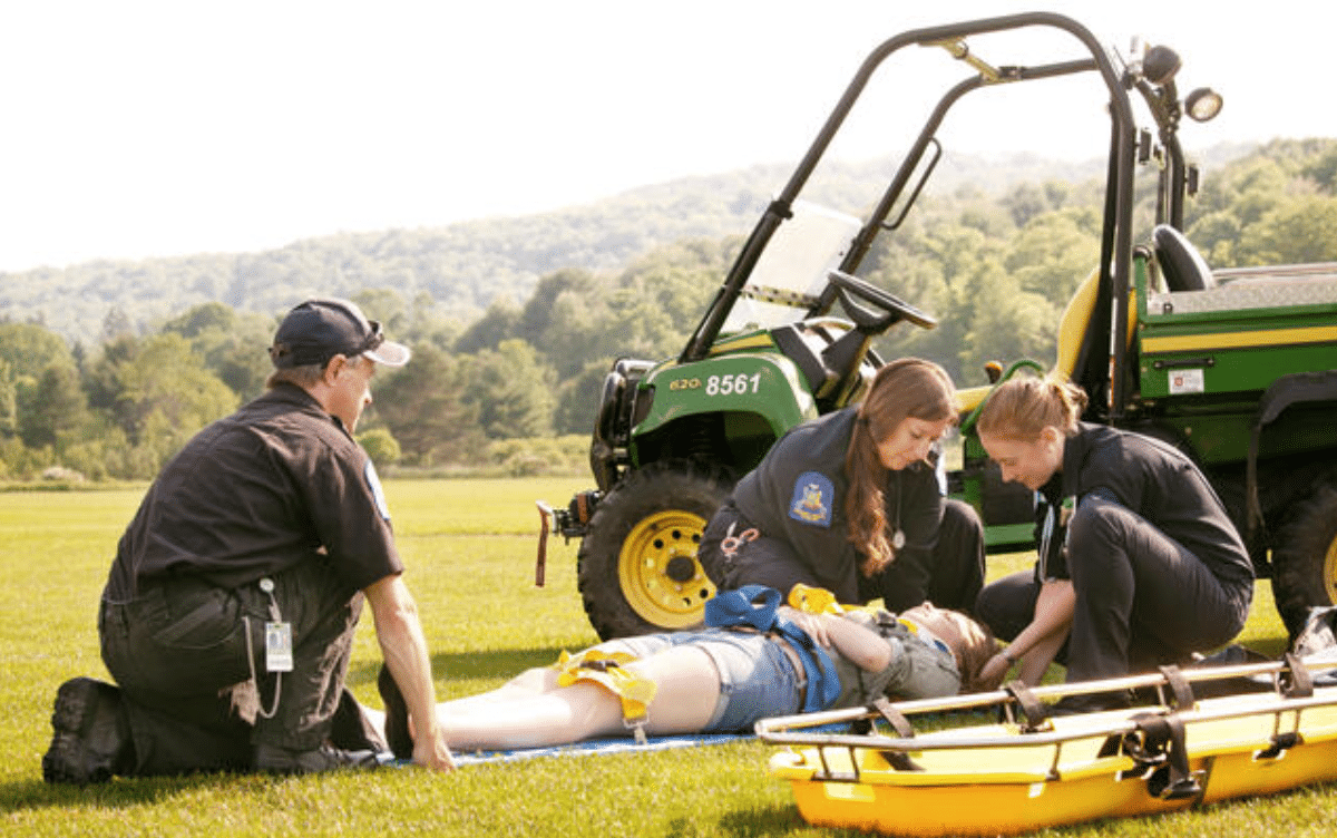 Three emergency responders attend to a person lying on grass beside a stretcher, bandaging their leg. A utility vehicle and trees are visible in the background.
